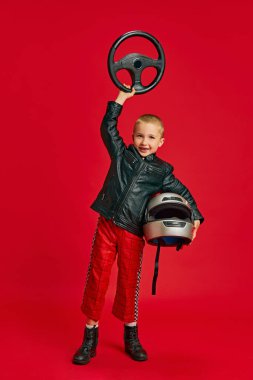 Coşkulu bir çocuğun uzun bir portresi, yarış kıyafeti giymiş küçük bir çocuk canlı kırmızı arka planda el kaldırıyor. Çocukların hayalleri, gelecekteki kariyerleri, meslekleri, oyunları, fantezileri. Ad