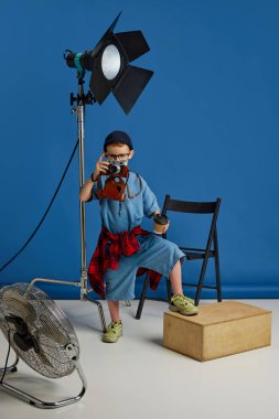 Kamera, kahve fincanı ve film setleriyle sıradan giyinmiş küçük bir çocuk mavi arka planda fotoğrafçı kariyeri hayal ediyor. Çocuk hayalleri, gelecekteki kariyer, oyunlar, fantezi. Ad
