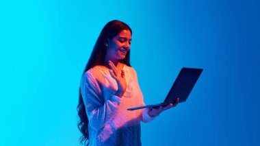 Orta Doğulu genç bir kadın mavi gradyan arka planda dizüstü bilgisayarında toplantı yaparken el sallıyor. Çevrimiçi alışveriş kavramı, eğitim ve uzaktan çalışma, teknoloji.