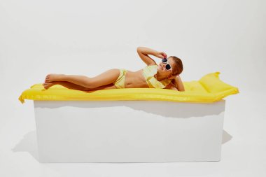 Küçük bir kız, mayo ve havalı gözlükler giymiş, canlı, güneşli sarı şişirilebilir bir yatakta uzanıyor ve beyaz stüdyo arka planında güneşleniyor. Sıcak yaz tatili kavramı, tatil, kamplar