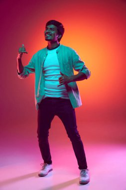 Neşeli genç Arap adam gündelik giysiler giyiyor ve neon ışıkta neon ışıkta dans edip dans ediyor. Gradyan stüdyo geçmişine karşı. Moda ve tarz kavramı, insan duyguları, müzik.