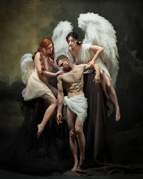 Stock image Two women with angelic wings support shirtless man wearing in white fabric, symbolizing psychological interplay between despair and hope. Concept of art, history, pain, vintage, beauty. Ad