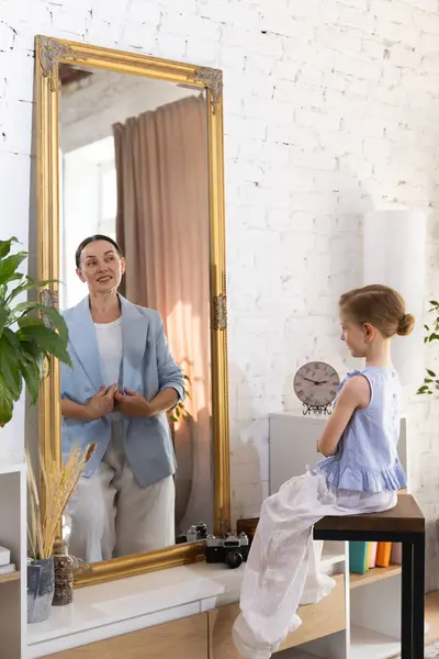 stock image Little girl sees her older self in mirror in blue elegant blazer, symbolizing evolution from childhood dreams to mature aspirations. Concept of past and future, generation, memories, fantasy.