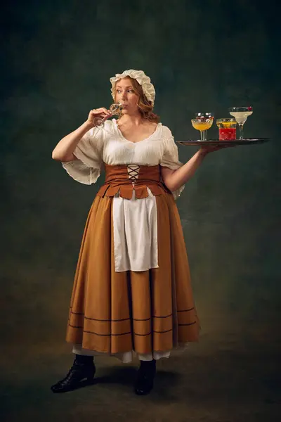 stock image Portrait of woman in historical attire, drinking cocktail and holding a tray with various alcoholic drinks, set against vintage green background. Concept of history, medieval bar, restaurant