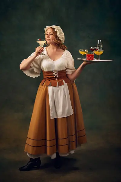 stock image Full-length portrait of woman in medieval clothes standing with tray of colorful cocktails, set against vintage green background. Bar poster, mixology. Concept of history, comparison of eras