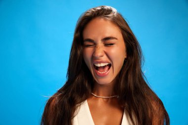 Duygusal, sinirli, sıradan giyinmiş genç bir kadın mavi stüdyo arka planına karşı bağırıyor. Stres, gerginlik. Moda renkleri. Olumsuz duygular, yüz ifadesi kavramı. Ad