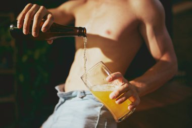 Barmen, gömleksiz genç adam ışık saçıyor, soğuk, bulanık arka planda bardağa bira döküyor. Alkolik içki. Yaz kavramı, içecek, parti, tatil, kutlama.