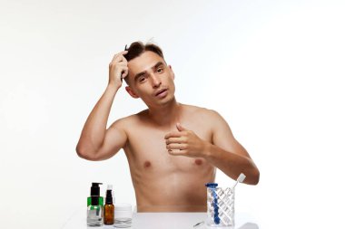 Çıplak gövdeli genç adam kameraya ayna gibi bakar ve saçını beyaz stüdyo arka planında tarar. Güzellik, özen, kozmetik, sağlık, erkeklik kavramı. Ad