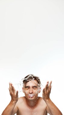 Poster. Yıkanan genç bir adam, modern banyoda beyaz stüdyo geçmişine karşı yüzüne su sıçratıyor. Cilt bakım rutini. Güzellik, özen, kozmetik, sağlık, erkeklik kavramı. Ad