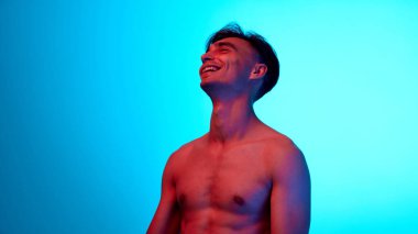 Genç adam üstsüz poz veriyor ve mavi arka plana karşı neon ışıkta gülüyor. Kırışıklıkları taklit ederek. Güzellik, bakım, kozmetoloji tedavisi, sağlık, erkeklik kavramı. Ad