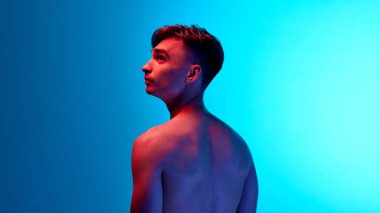 Üstsüz bir adamın arka mücadelesi, neon ışıkta yukarı doğru dramatik bir poz vererek kaslı vücudunu mavi eğimli arka planına vurguluyor. Güzellik, bakım, kozmetoloji, sağlık, erkeklik kavramı.