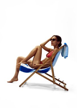 Güverte sandalyesinde uzanan, kırmızı bikini giyen ve beyaz stüdyo arka planında güneş gözlüğü takan kadınlar. Güzellik kavramı, vücut bakımı, cilt bakımı, yaz mevsimi, koruma.