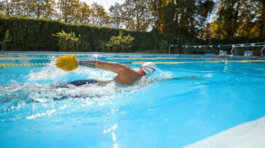 Beyaz yüzme şapkası takan atletik bir adam ve güneş havuzundaki performansını artırmak için sarı raket kullanan gözlükler serbest yüzer. Profesyonel spor anlayışı, aktif ve sağlıklı yaşam tarzı. Ad