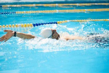 Formda bir adam, yüzücü, beyaz şapkalı, havuzda ileri doğru itilmiş, kolu açık havuzda serbest stil vuruşuyla güçlü bir şekilde uzanmış. Spor anlayışı, rekabet, aktif ve sağlıklı yaşam tarzı. Ad
