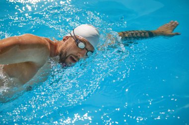 Felce odaklanmış, beyaz şapkalı bir yüzücü havuza giriyor, kolu uzanıyor ve güneş ışığı su yüzeyinden yansıyor. Profesyonel spor, rekabet, aktif ve sağlıklı yaşam tarzı kavramı.