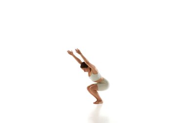 Atletik giyinen bir kadının dinamik fotoğrafı sandalye pozu veriyor, dizlerini büküyor ve beyaz stüdyo geçmişine yaslanıyor. Yoga. Güzellik, farkındalık ve denge anlayışı, akıl sağlığı, konsantrasyon. Ad