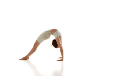 Atletik giyinen bir kadın, sırtını esneterek ve beyaz stüdyo geçmişine karşı esneklik göstererek tam bir yoga pozisyonu sergiler. Güzellik, farkındalık ve denge kavramı, akıl sağlığı. Ad