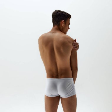 Beyaz donlu, formda bir gencin arka plan beyaz stüdyo arka planında bir eli omzunda durduğu arka plan fotoğrafı. Vücut bakımı, erkek sağlığı, doğal güzellik, erkeklik kavramı. Ad