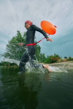 Dalgıç kıyafetli ve yüzme şapkalı bir sporcu göle turuncu şamandıra ile su sıçratır ve yemyeşil ortamda yüzmeye hazırdır. Spor müsabakası, spor, eğlence, sağlıklı yaşam tarzı kavramı.