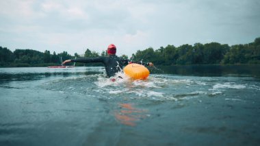 Kırmızı şapkalı ve siyah dalış kıyafetli bir yüzücü, turuncu şamandırayla donatılmış, dingin göle giriyor, su etrafa sıçrıyor. Spor müsabakası, spor, eğlence, sağlıklı yaşam tarzı kavramı.