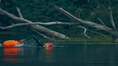 Dalgıç kıyafetli triatloncu, doğal yeşilliklerle çevrili sakin gölde turuncu şamandıra eşliğinde açık denizde yüzme pratiği yapıyor. Spor müsabakası, spor, eğlence, sağlıklı yaşam tarzı kavramı.