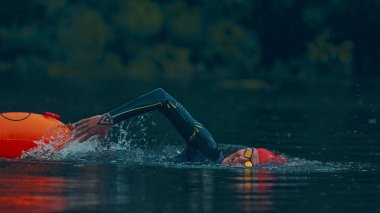 Kırmızı şapkalı ve siyah dalgıç kıyafetli triatloncu dingin göl boyunca ormanın arka planında turuncu şamandırayı çekiyor. Spor müsabakası, spor, eğlence, sağlıklı yaşam tarzı kavramı.