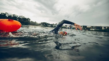 Siyah dalgıç kıyafetli ve kırmızı şapkalı sporcu suda hareket ediyor, kolları gölü kesiyor, triatlon eğitimini yakalıyor. Spor müsabakası, antrenman, eğlence anlayışı