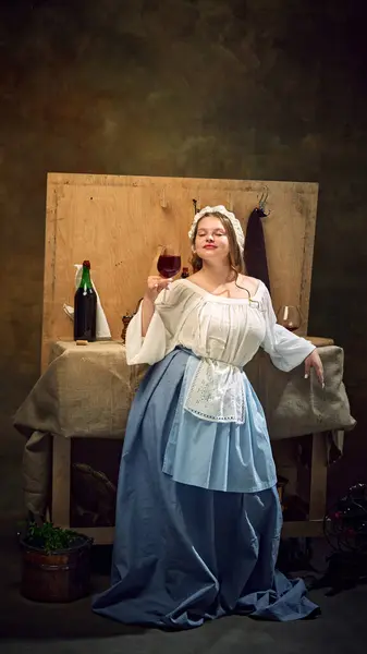 stock image Joyful, young woman in old-fashion dress holding glass of wine standing leaned on bar counter at pub against vintage background. Concept of bar, pub, Bavarian style, Oktoberfest. Comparisons of eras.