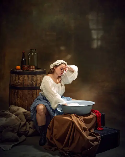 stock image Medieval woman in traditional attire sits wearily by basin, washing clothes, with barrels and simple household items surrounding her. Concept of Bavarian style, Oktoberfest. Comparisons of eras.