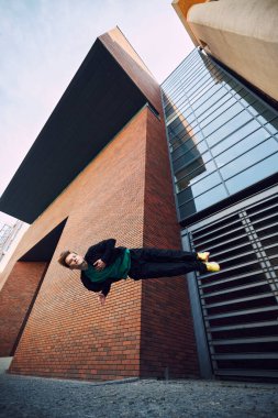 Yeşil gömlekli ve turuncu şapkalı break dansçı etkileyici bir salto sergiliyor, yerçekimine stille meydan okuyor. Şehir yaşam tarzı. Ekstrem spor anlayışı, serbest stil, aktivite, hareket. Ad