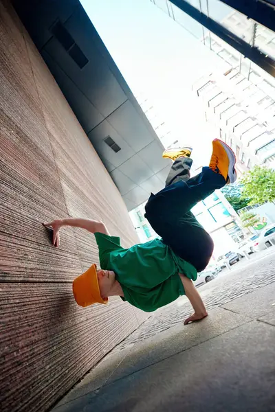 stock image Breakdancer in green shirt and orange bucket hat strikes gravity-defying pose, balancing on one hand against city wall, shows agility and flair. Concept of lifestyle, extreme sport, freestyle. Ad