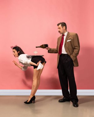 Yüksek topuklu giyen kadın sırtında tepsiyle eğiliyor, takım elbiseli adam pembe arka plana karşı şişeden su dökerken. Güzellik anlayışı, satış, moda ve tarz, klasik. Ad