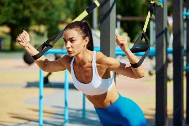 Kadın TRX egzersizi yapıyor, vücudu öne eğiliyor, kolları açık. Sporcu antrenmanla meşgul. Spor salonu açık havada. Profesyonel spor, vücut geliştirme, moda ve tarz kavramı. Ad