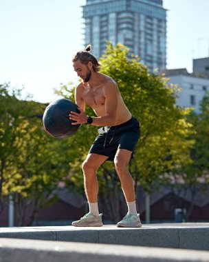 Güneşli bir günde kaslı, sportif vücut eğitimi almış üstsüz genç bir adam tıp topuyla yoğun egzersizler yapıyor. Spor anlayışı, aktif ve sağlıklı yaşam tarzı, fitness