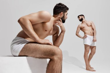 İki beyaz iç çamaşırlı adam düşünceli ve yoğun bakışlar atıyorlar, biri oturuyor ve diğeri sıcak gri arka planda duruyor. Erkek sağlığı, güzellik ve moda, fitness, erkeklik kavramı.