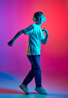 Küçük çocuk günlük kıyafetlerini giymiş, kulaklıkla dans ediyor, mavi-pembe renkli gradyan stüdyo arka planına karşı. Çocuk kavramı, insani duygular, moda ve tarz, yaşam tarzı. Ad