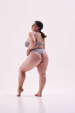 Kendine güvenen, güzel bir kadın, gri iç çamaşırlarıyla poz veren bir model, kalçaları beyaz stüdyo arka planında. Doğal güzellik kavramı, vücut pozitifliği, kendini sevme, kaplıca tedavisi.