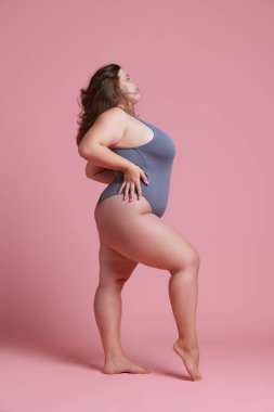 Pembe stüdyo arka planına karşı gri mayo giymiş genç ve tombul bir kadının yan görünümü. Doğal güzellik kavramı, vücut pozitifliği, kendini sevme, kaplıca tedavisi. Ad