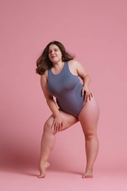 Gri mayo giymiş genç bir kadın, kendine güvenen ve kendini beğenmişlik taslayan pembe stüdyo geçmişine karşı gururlu bir poz veren. Doğal güzellik kavramı, vücut pozitifliği, kendini sevme, kaplıca tedavisi. Ad