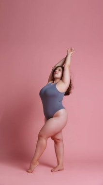 Güçlü bir kadın, artı beden modeli, forma uygun bir vücut giysisi içinde, pembe stüdyo arka planına karşı el kaldırıyor. Doğal güzellik kavramı, vücut pozitifliği, kendini sevme, kaplıca tedavisi. Ad
