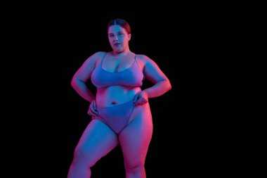 Kendine güvenen bir kadın, iç çamaşırı giyen bir model, siyah stüdyo arka planında parıldayan bir ışık. Doğal güzellik kavramı, vücut pozitifliği, kendini sevme, kaplıca tedavisi. Ad