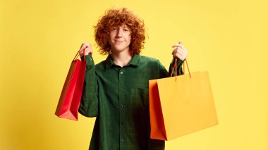 Zevkli adam portresi, kıvırcık saçlı öğrenci, sarı arka planda hediyeler taşıyan. Kara Cuma kavramı, Siber Pazartesi, moda, hızlı satış. Ad