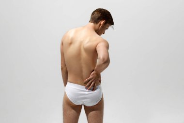 Beyaz şortlu genç adamın arka görünüşü, eli belinde dinleniyor, rahatsızlık ya da tefekkür izlenimi veriyor. Erkek sağlığı kavramı, doğal güzellik insanları, moda, kişisel bakım.