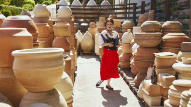 Geleneksel kıyafetli bir kadın, elinde bir bardak şarapla, sıcak güneş ışığı altında çömleklerin arasında otururken gülümseyerek onu kaldırıyor. Yaz sonu kavramı, hasat, seyahat, doğal güzellik, kültür.