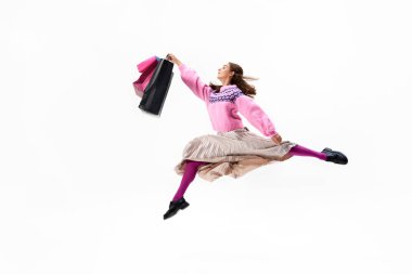 Elinde çantalarla havaya sıçrayan genç kadın, parlak ve şık kıyafetlerini beyaz stüdyo geçmişine karşı sergiliyor. Alışveriş kavramı, Kara Cuma, moda ve tarz, satış sezonu.