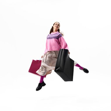 Pembe kazaklı kadın, beyaz stüdyo arka planında renkli alışveriş torbaları taşıyarak sevinçle havada zıplıyor. Alışveriş anlayışı, Kara Cuma, moda ve tarz, satış sezonu. Ad
