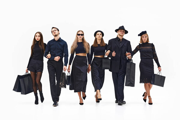 Группа модных людей в полностью черных нарядах уверенно идет вперед с сумками в руках, выглядя уравновешенно и шикарно. Концепция покупок, Черная пятница, мода и стиль, сезон продаж. Объявление