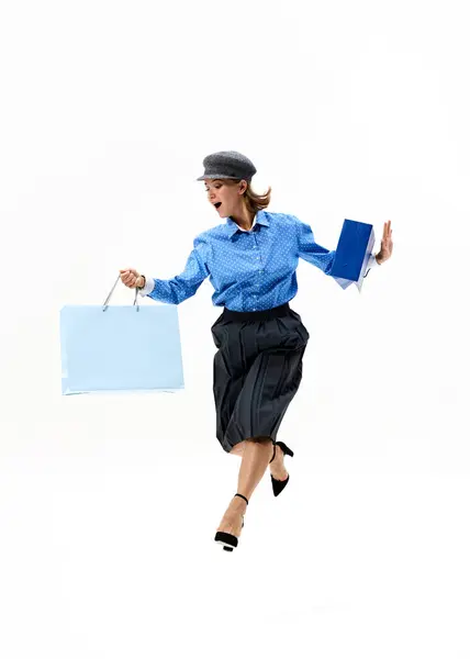 stock image Leaping in joy with bags in hand, woman in blue polka-dot shirt and black skirt celebrates her shopping triumph against white studio background. Concept of shopping, Black Friday, fashion and style