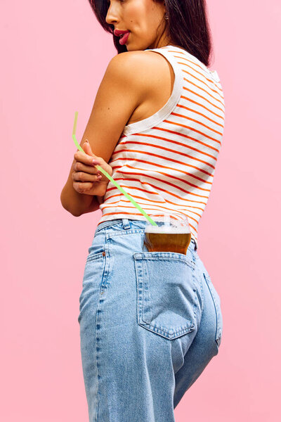 Stylish woman drinks from glass in her back pocket with straw, against pink background, showing off quirky and carefree attitude. Concept of holidays, celebration, festive mood. Pop art style.