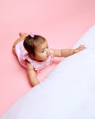 Bebek pembe desenli giyinmiş, yumuşak beyaz yastığa uzanıyor, pembe arka planda tuhaf bir ifade var. Güzellik, ebeveynlik, çocukluk, yaşam ve doğum kavramı.